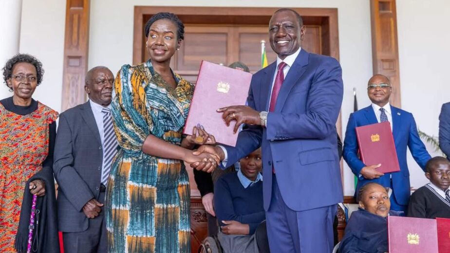 Kenya’s President William Ruto hands over the Persons with Disabilities Act 2025 to Senator Crystal Asige, who is visually impaired, after signing the bill into law at State House, with officials and persons with disability in the background.