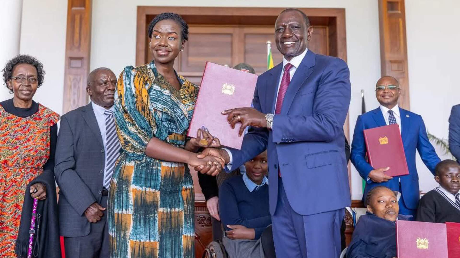 Kenya’s President William Ruto hands over the Persons with Disabilities Act 2025 to Senator Crystal Asige, who is visually impaired, after signing the bill into law at State House, with officials and persons with disability in the background.