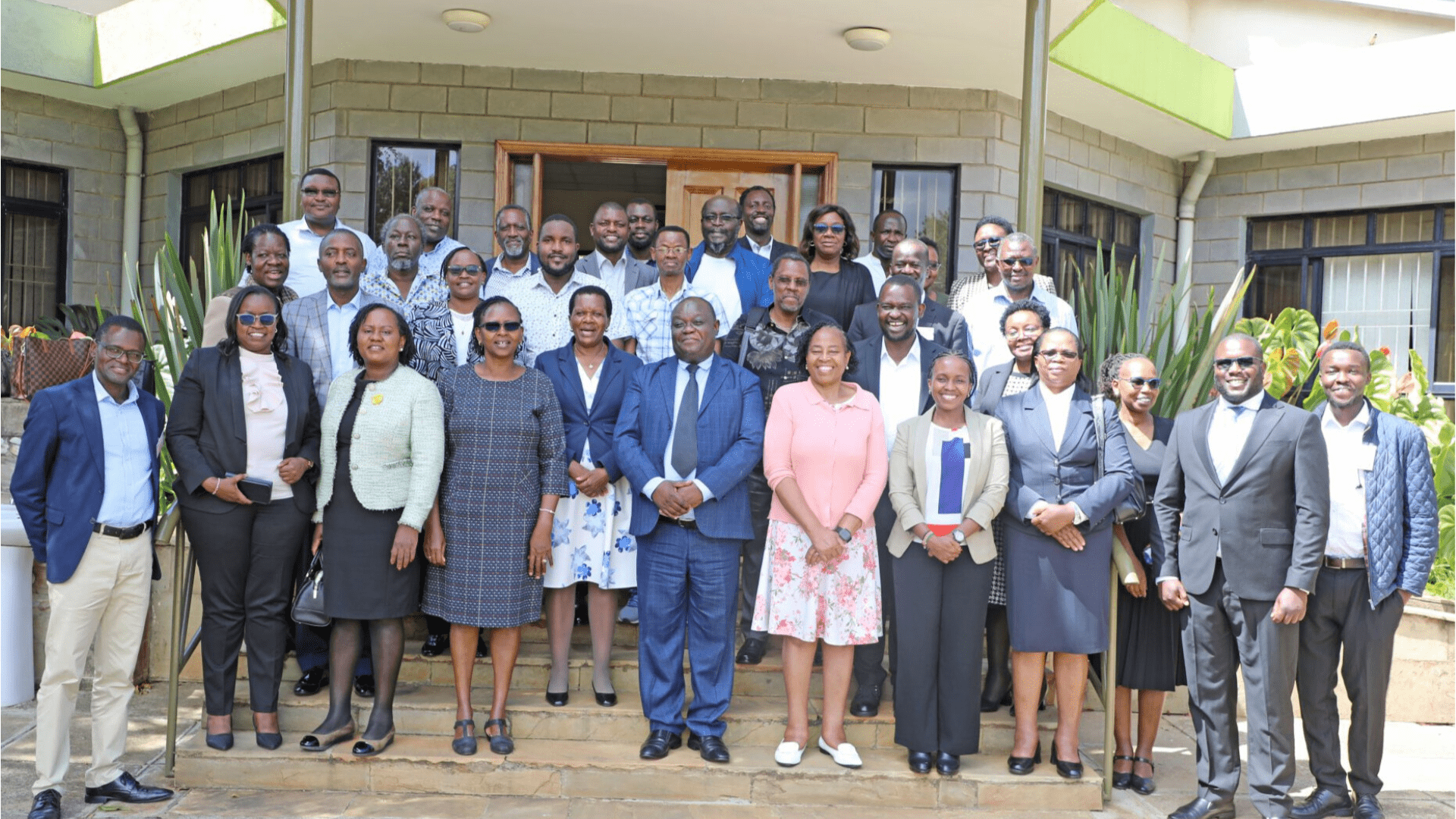 A group photo of around 40 trainees and instructors standing on the steps of a building entrance at the Kenya School of Government in Nairobi. They are dressed in business casual and formal attire, posing together outdoors with smiles, plants, and a brick wall backdrop.