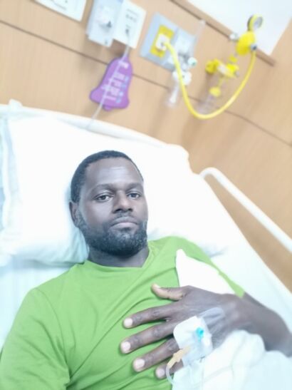 Kizito Namulanda in a green T-shirt lying in a hospital bed at Max Hospital during cancer treatment, with an IV line in his hand