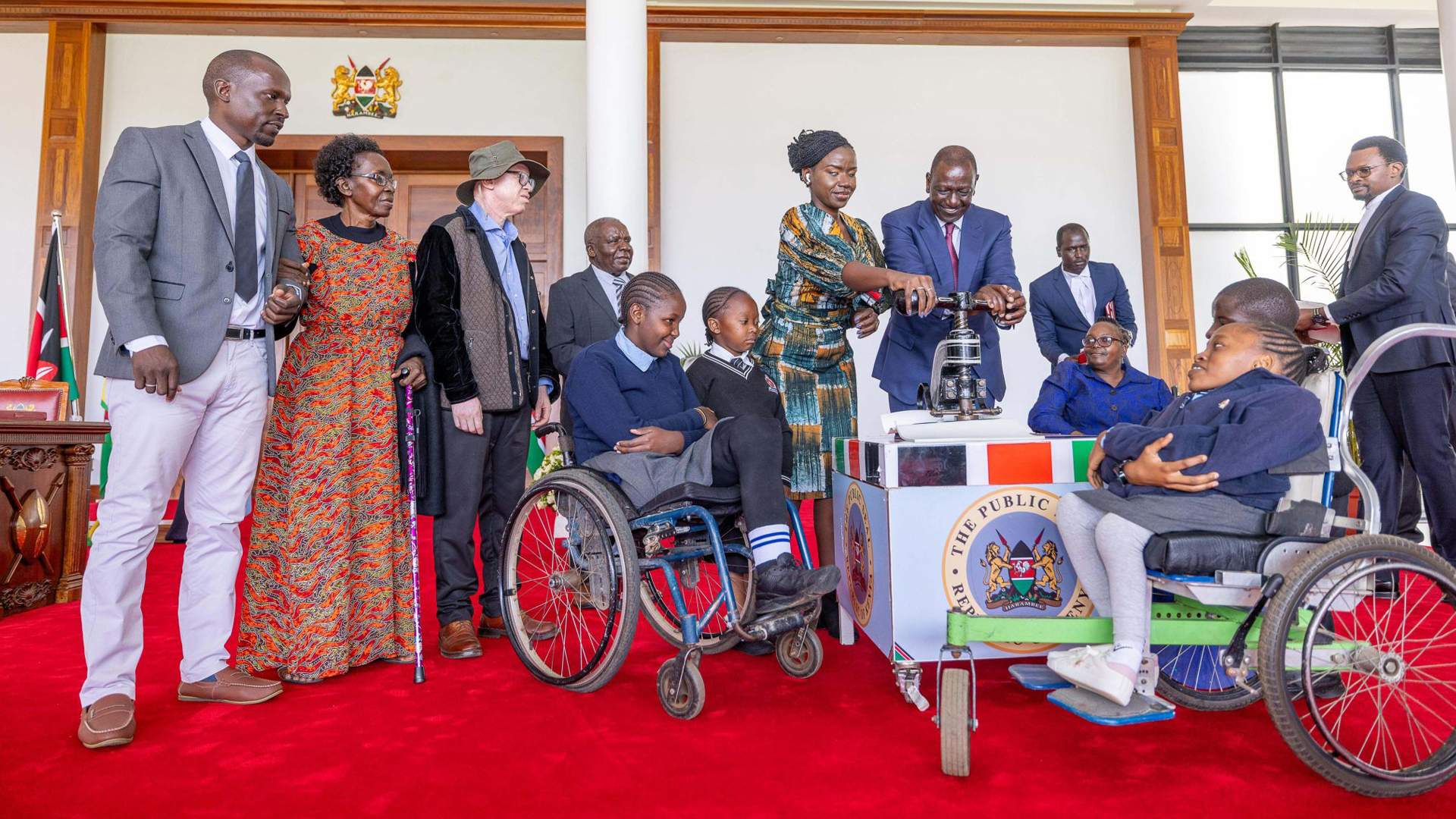 Kenya’s President William Ruto and Nominated Senator Crystal Asige (visually impaired) seal the Persons With Disability Act 2025 at State House, Nairobi, while persons with disabilities look on; a seal is placed on a document beside a ballot-like device, with a group of officials and attendees in the background.