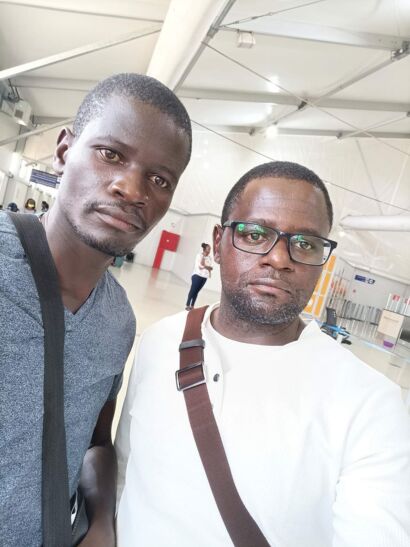 A photo of two men standing close together in an airport terminal. The man on the left wears a gray shirt and has a strap over his shoulder; the man on the right wears glasses and a white shirt with a brown shoulder bag. They pose for a selfie, with an airy, indoor backdrop and other travelers in the background. The caption notes that this is Kizito Namulanda (right) with his brother Peter Musumba traveling to New Delhi in April 2022.
