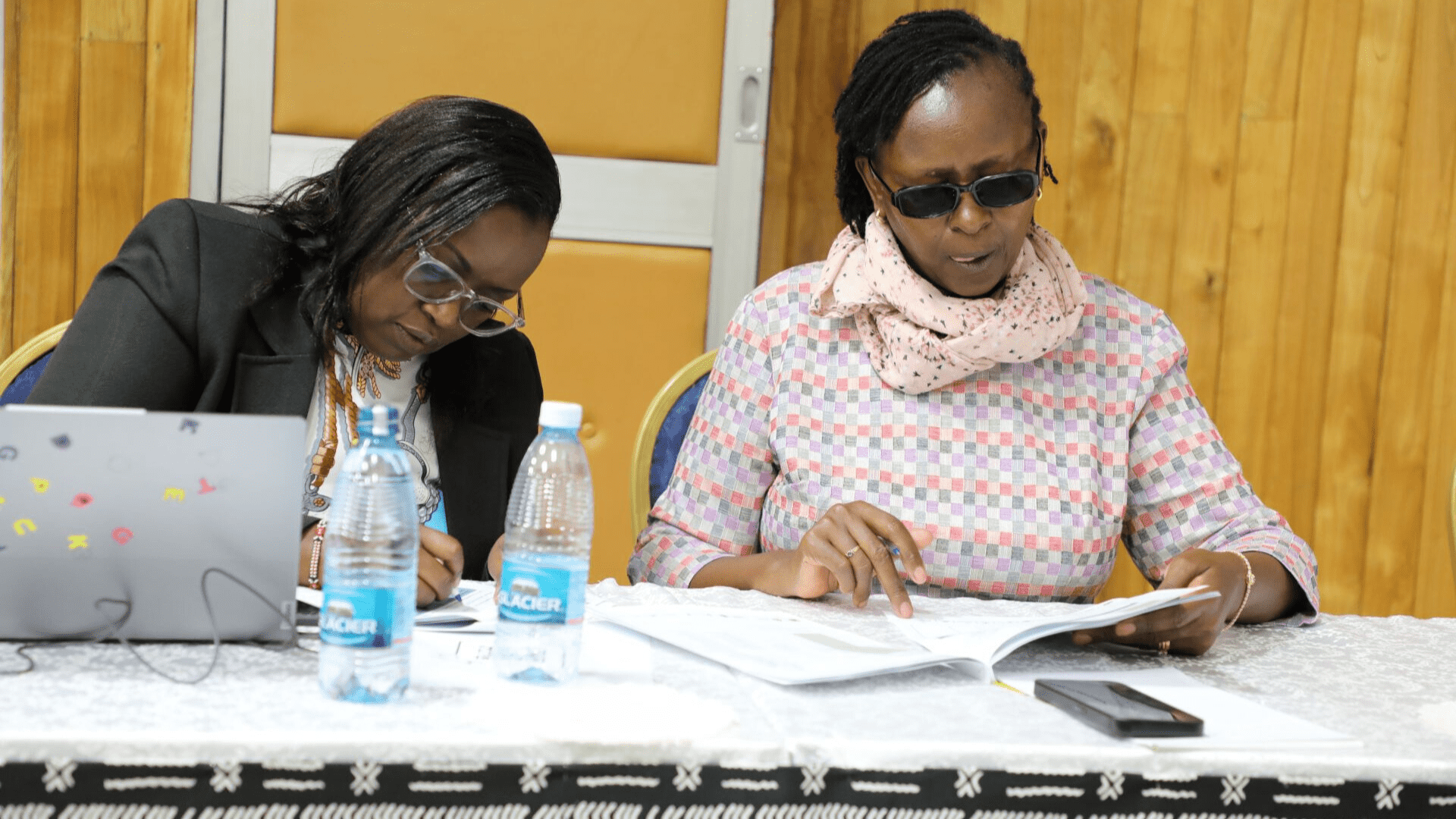 Two women seated at a conference table during the ELRC Digital and ICT Accessibility training at the Kenya School of Government in Nairobi. Left is Hon. Lady Justice Stella Rutto and right is Hon. Lady Justice Monica Mbaru; both are reviewing documents with bottles of water and laptops visible in front of them, other attendees in the background. 