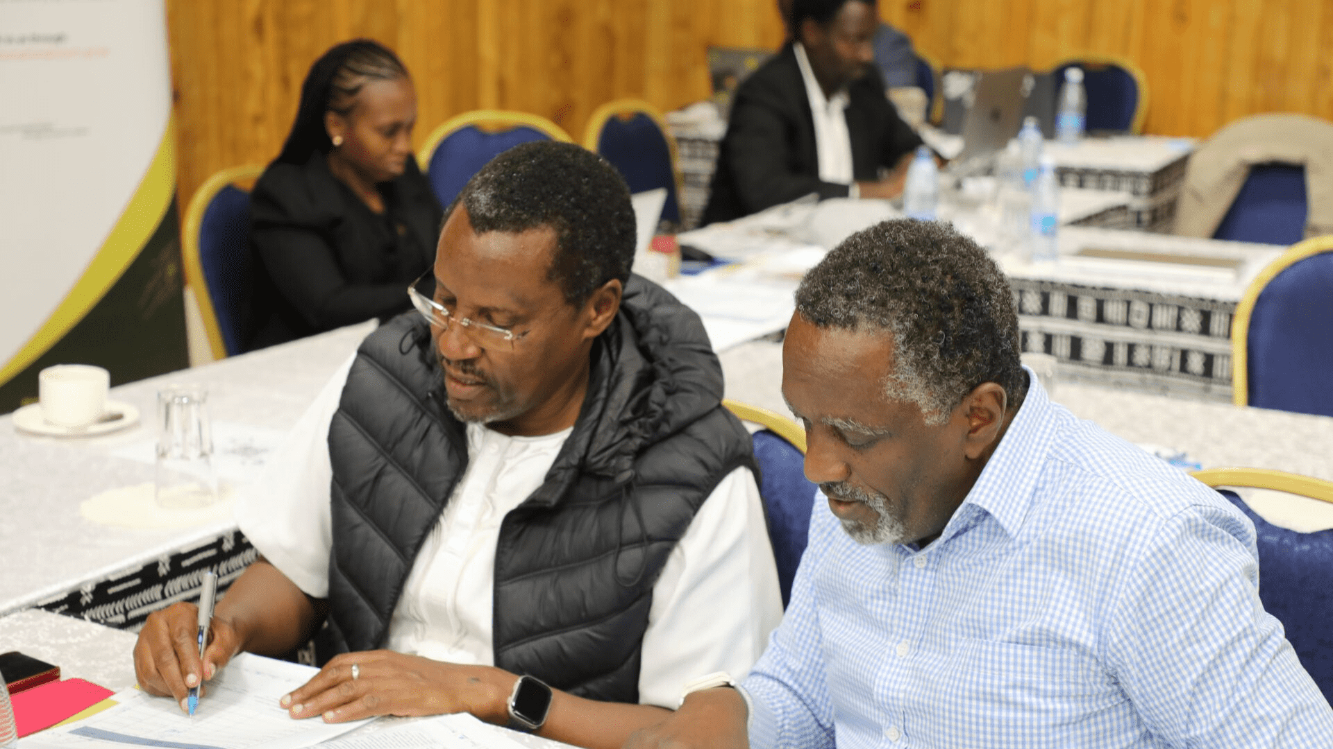 Two men seated at a conference table during the ELRC Digital and ICT Accessibility training at the Kenya School of Government in Nairobi. Left is Hon. Mr. Justice Jacob Gakeri Kariuki and right is Hon. Mr. Nzioki wa Makau; both appear engaged in documents with other attendees in the background. 